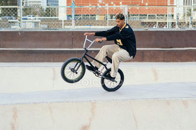 Vista lateral del joven deportista afroamericano realizando trucos en bicicleta de prueba en el parque de skate de la ciudad - foto de stock