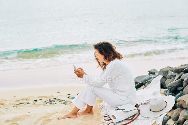 Side view of tranquil female sitting on beach near sea and enjoying music on earphones and using mobile — Foto stock