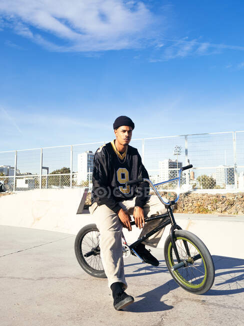 Молодий етнічний хіпстер чоловік-спортсмен в холодному одязі сидить на велосипеді BMX, дивлячись далеко в місто в сонячний день — стокове фото