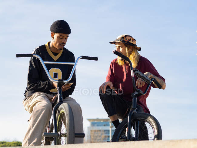 Desde abajo de jóvenes deportistas multiétnicos alegres en bicicleta de prueba mirándose durante el saludo en la ciudad - foto de stock