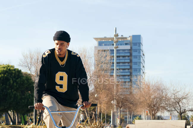 Young ethnic hipster male athlete in cool wear sitting on BMX bike while looking away in town on sunny day — Fotografia de Stock