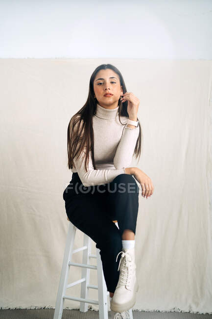 Jovem sem emoção e pensativa mulher olhando para a câmera sentada em um banquinho no fundo do estúdio durante o dia — Fotografia de Stock