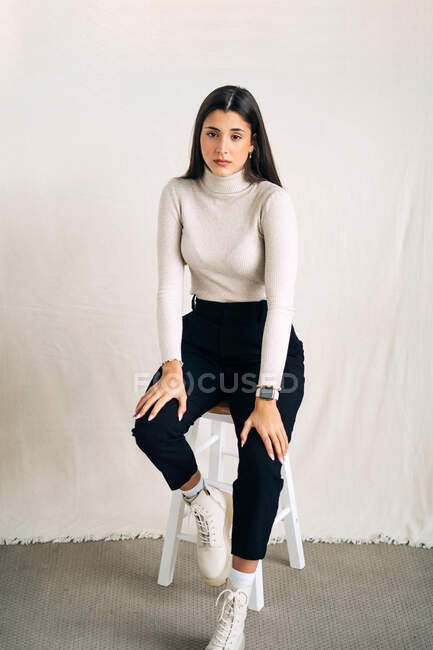 Unbewegte junge, nachdenkliche Frau blickt tagsüber in die Kamera, die auf einem Hocker im Studiohintergrund sitzt — Stockfoto