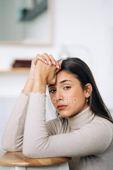 Неемоційна молода вдумлива жінка, дивлячись на камеру, що спирається на табурет на студійному фоні вдень — стокове фото