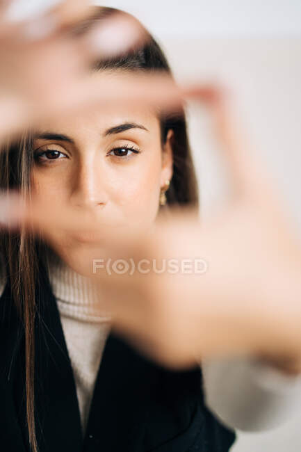 Молода роздумна жінка в повсякденному одязі робить фоторамку з пальцями і дивиться на камеру на світлому фоні — стокове фото