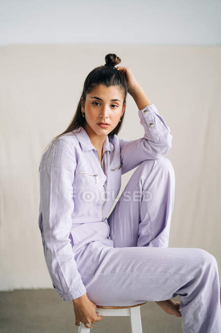 Неемоційна молода вдумлива жінка в стильному загальному вигляді дивиться на камеру, сидячи на табуреті на студійному фоні вдень — стокове фото