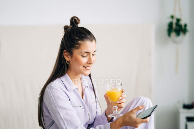 Jovem fêmea amigável com vidro de suco de laranja navegando telefone celular em casa — Fotografia de Stock