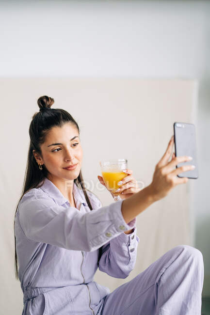 Jovem fêmea amigável com vidro de suco de laranja tomando auto retrato no celular em casa — Fotografia de Stock