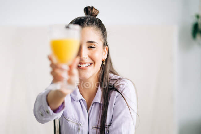 Молодая веселая женщина закрывает глаза стаканом вкусного напитка, глядя в камеру дома — стоковое фото