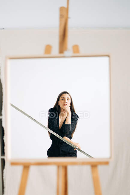 Junge nachdenkliche Frau in schwarzer Kleidung und Ohrring blickt in die Kamera im Spiegel auf Staffelei gestellt — Stockfoto
