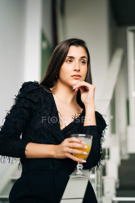 Young thoughtful female in black outfit with glass of orange juice touching chin while looking away on staircase in building — Stock Photo