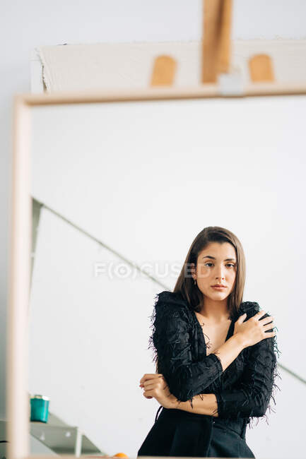 Jovem mulher pensativa em desgaste preto e brinco olhando para a câmera no espelho colocado no cavalete — Fotografia de Stock