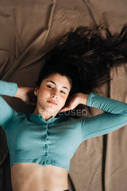 Visão superior da jovem mulher consciente olhando para a câmera deitada na cama com têxteis amassados em casa — Fotografia de Stock