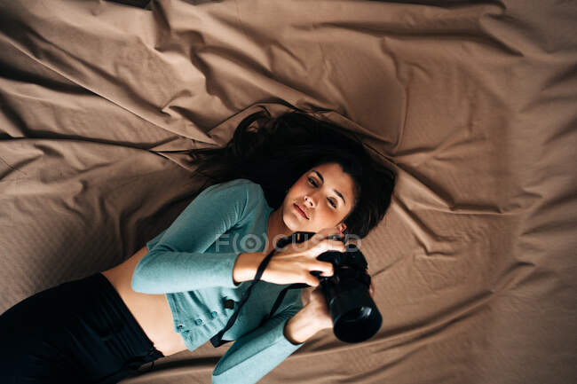 Overhead view of millennial female with professional photo camera lying on crumpled textile and looking at camera at home — Stock Photo