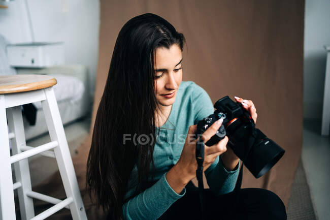 Femme millénaire avec appareil photo professionnel assis sur fond textile froissé brun à la maison — Photo de stock