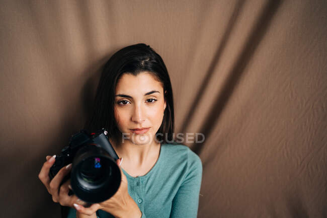 Millennial-Weibchen mit professioneller Fotokamera sitzt auf braunem, zerknittertem Textilhintergrund und schaut zu Hause in die Kamera — Stockfoto