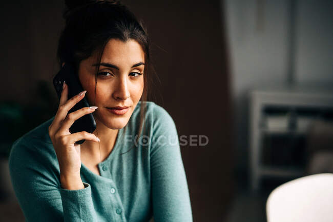 Jeune femme charmante parlant sur un téléphone portable tout en regardant la caméra à la maison sur fond flou — Photo de stock