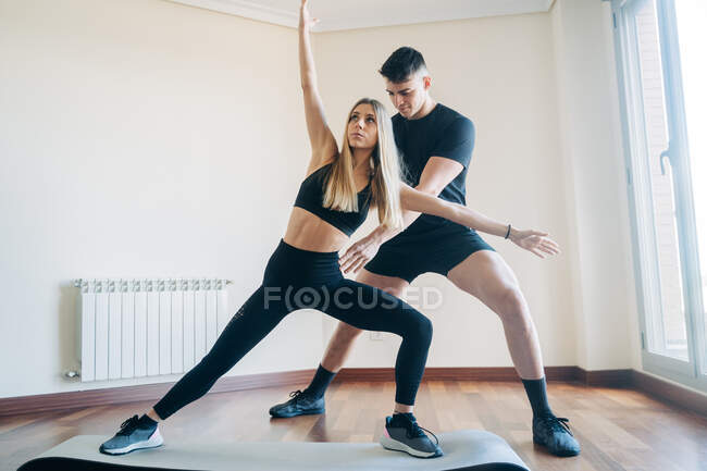 Full body of male personal instructor supporting woman doing lunges exercise with raised arms on mat during workout at home — Stock Photo