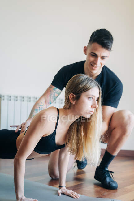 Full body of male personal instructor supporting woman doing lunges exercise with raised arms on mat during workout at home — Stock Photo