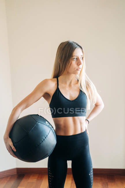 Athletic female with abs wearing top and leggings standing with heavy black ball for training in light room during workout — Stock Photo