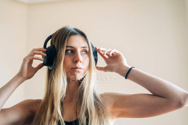 Зосереджена жінка з довгим волоссям в навушниках слухає музику і дивиться в сторону, стоячи в світлій кімнаті з руками біля голови — стокове фото