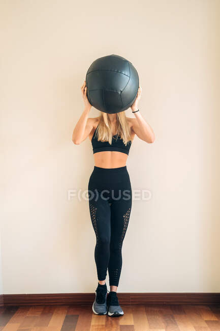 Спортивная женщина с прессом и леггинсами, стоящая с тяжелым черным мячом для тренировки в легкой комнате во время тренировки — стоковое фото