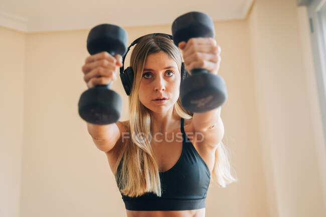 Entschlossene Frau in Sportbekleidung mit Kopfhörern, die Musik hört, während sie zu Hause Ausfallübungen mit Gewicht macht — Stockfoto
