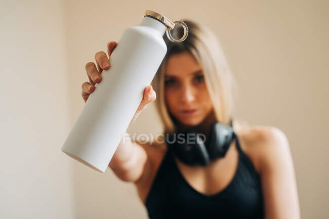 Mulher calma com fones de ouvido vestindo activewear olhando para a câmera e beber água enquanto descansa no quarto após o treinamento em casa — Fotografia de Stock