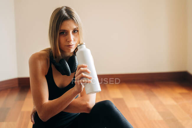 Спокойная женщина с наушниками в активной одежде, смотрящая на камеру и питьевую воду во время отдыха в комнате после тренировки дома — стоковое фото
