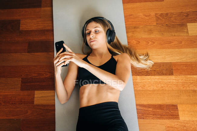 Visão superior da fêmea em fones de ouvido ouvindo música e surfando celular enquanto deitado no tapete após o treino em casa — Fotografia de Stock