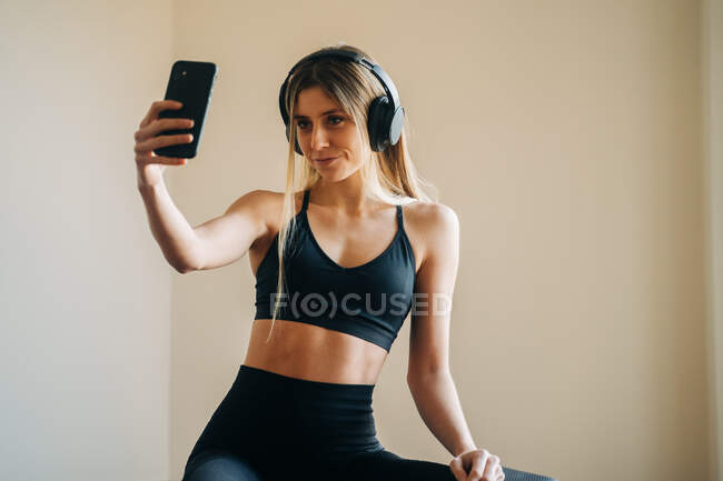 Sportliche Frau in Aktivkleidung mit Kopfhörer, Musik hörend und Selbstporträt auf dem Smartphone, während sie nach dem Training im Zimmer sitzt — Stockfoto
