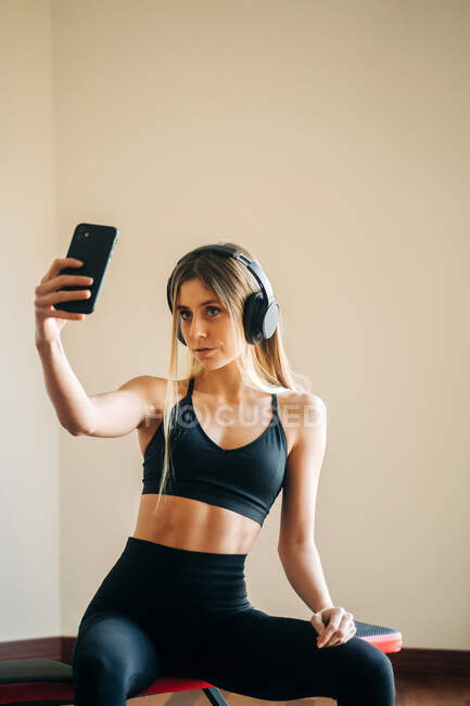 Спортивна жінка в активному одязі з навушниками, що слухають музику і знімають самопортрет на смартфоні, сидячи в кімнаті після тренування — стокове фото