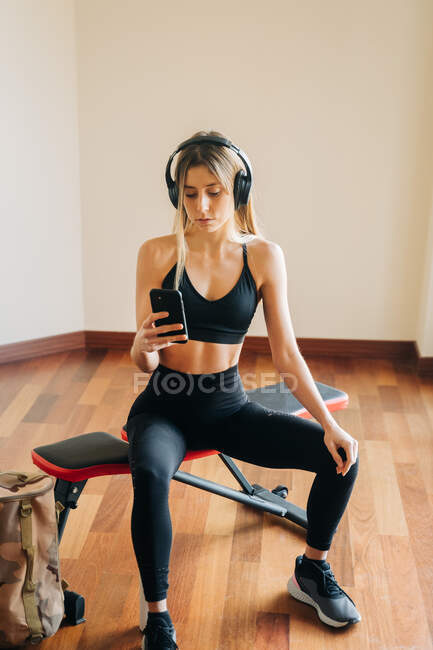 Спортивная женщина в спортивной одежде с наушниками, слушающая музыку и делающая автопортрет на смартфоне, сидя в комнате после тренировки — стоковое фото