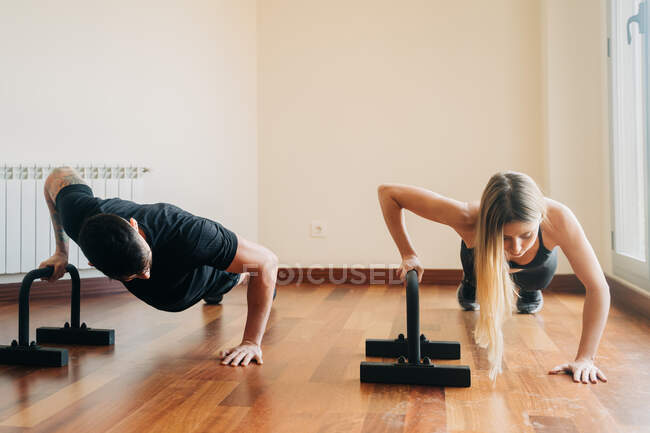 Determinado hombre y mujer practicando ejercicio usando flexiones de acero se para mientras construye músculos del pecho durante el entrenamiento en la habitación en casa - foto de stock