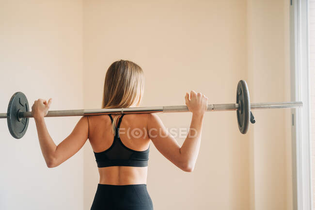 Mujer activa fuerte que usa un atuendo deportivo negro mirando a la cámara mientras hace sentadilla en la espalda durante el entrenamiento en la sala de luz - foto de stock