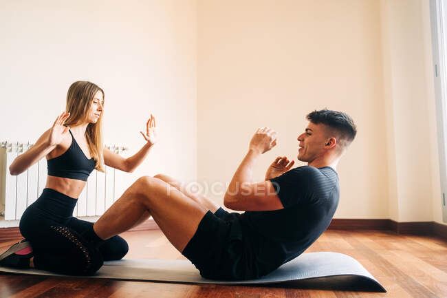 Vista lateral de la mujer en forma con las manos levantadas ayudando al hombre deportivo mientras practica crujidos en la estera en la habitación en casa - foto de stock
