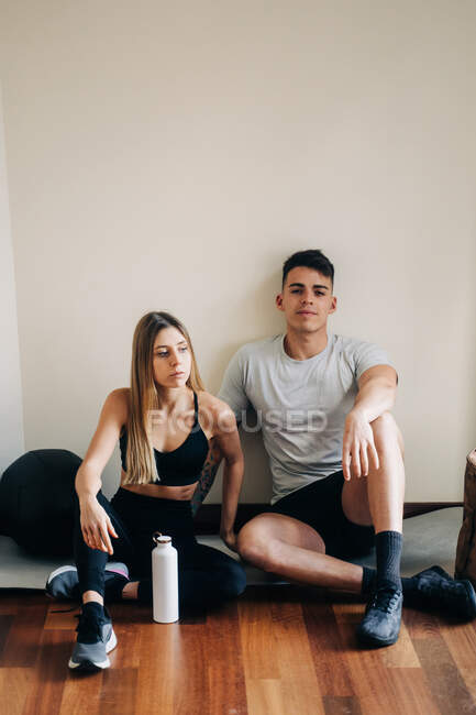 Ganzkörper Mann und Frau in sportlicher Kleidung sitzen nach dem Training mit überkreuzten Beinen auf Parkett in Wandnähe mit einer Flasche Wasser — Stockfoto