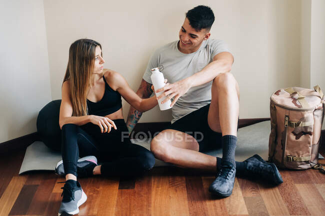Full body of man and woman in sportive wear sitting with crossed legs on parquet near wall with bottle of water after workout — Stock Photo