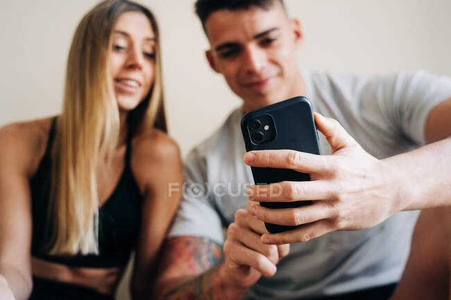 Conteúdo homem e mulher vestindo celular surfe sportswear enquanto sentado no chão cruzou a perna perto da parede com garrafa de água — Fotografia de Stock