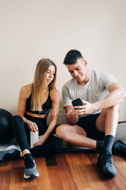 Content man and woman wearing sportswear surfing cellphone while sitting on floor crossed legged near wall with bottle of water — Stock Photo