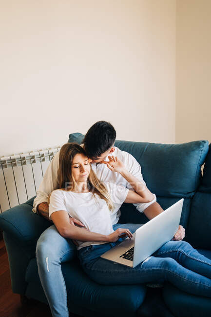 From above of tender boyfriend and focused girlfriend browsing netbook while lying on sofa and caressing on sofa near wall in living room — Stock Photo
