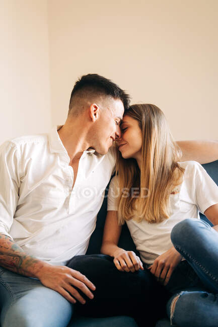 Side view of romantic boyfriend and girlfriend touching foreheads while caressing each other on couch in living room at home — Stock Photo