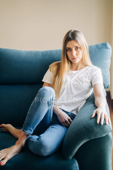 Pieno corpo di calma a piedi nudi femminile in jeans guardando la fotocamera mentre seduto sul divano con gambe incrociate in stanza luce — Foto stock