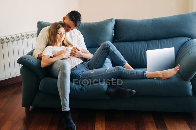 Сверху нежный бойфренд и целенаправленная девушка просматривает нетбук, лежа на диване и лаская на диване возле стены в гостиной — стоковое фото