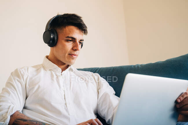Konzentrierter Mann mit Kopfhörer hört Musik und surft modernes Netbook, während er zu Hause auf der Couch sitzt — Stockfoto