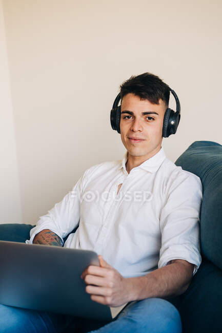 Сконцентрированный мужчина в наушниках, слушающий музыку и занимающийся серфингом в современном нетбуке, сидя на диване в гостиной — стоковое фото