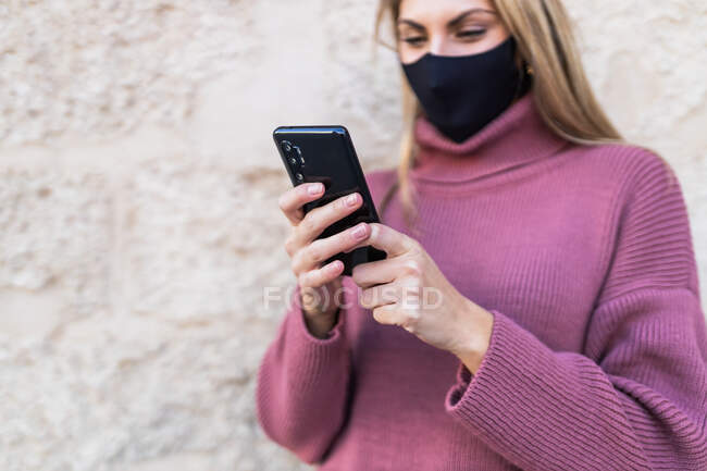 Lächelnde Frau in Schutzmaske surft mit Handy im Internet, während sie auf der Straße gegen Hauswand steht — Stockfoto