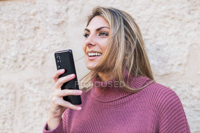 Tender charming female standing in city street with smartphone recording a voice near stone old wall looking away — Stock Photo