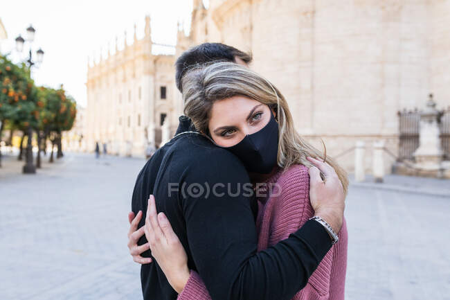 Vista lateral de la mujer cariñosa en máscara abrazando tiernamente hombre irreconocible mientras está de pie en la ciudad durante el paseo en fin de semana - foto de stock