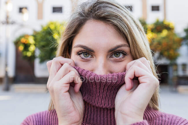 Tender charming female covering face with warm knitted sweater while standing in city street and looking at camera — Stock Photo